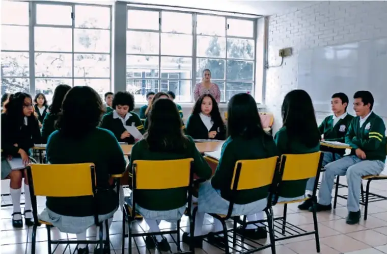 Fotografía de estudiantes de secundaria sentados en círculo en un aula con ventanales, participando en una asamblea o reunión de trabajo colaborativo; una docente observa de pie al fondo.