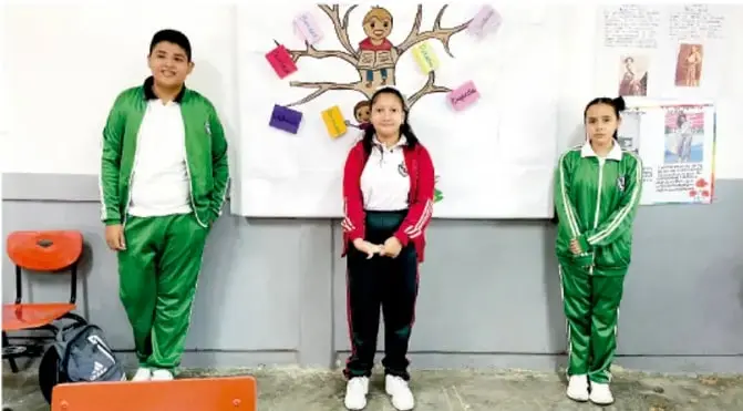 Fotografía de tres estudiantes con uniforme deportivo verde frente a un mural escolar con un árbol decorado, presentando su proyecto de cultura de paz.