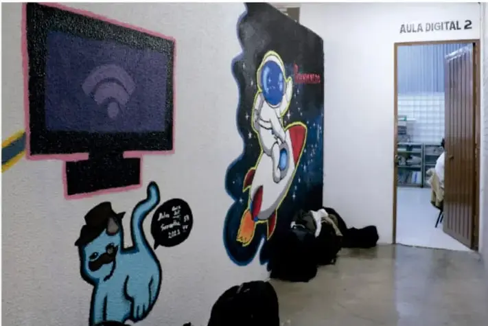 Fotografía de un mural pintado en la pared de un pasillo escolar: a la izquierda un monitor con señal inalámbrica y debajo un gato azul con boina; a la derecha un astronauta en un cohete sobre fondo oscuro. Al fondo, una puerta con el rótulo "Aula Digital 2" y objetos negros en el piso.