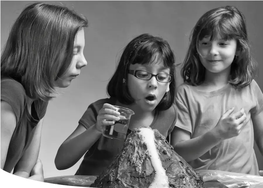 Fotografía en blanco y negro de tres niñas observando con sorpresa un modelo de volcán con erupción de espuma; la niña del centro sostiene un vaso con líquido.