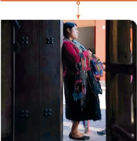 Mujer indígena con ropa tradicional cargando un rebozo, de pie junto a una puerta.