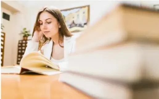 Fotografía de una joven leyendo un libro junto a una pila de volúmenes, ilustrando la lectura como parte de la formación del ensayista.