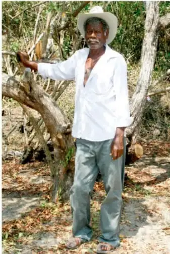 Fotografía de un hombre afrodescendiente con sombrero de paja y camisa blanca, apoyado en un árbol en un entorno rural con vegetación seca.