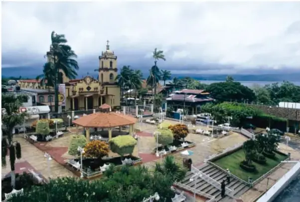 Plaza principal de un pueblo (Catemaco) con quiosco de techo anaranjado, iglesia con torres y palmeras, bajo cielo nublado; al fondo se alcanza la laguna.