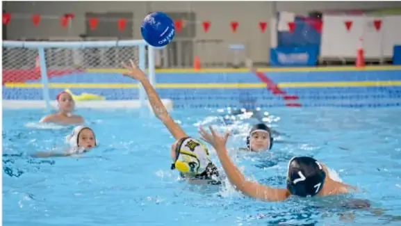 Fotografía de un partido de waterpolo en piscina: una jugadora lanza el balón azul hacia el arco mientras otras jugadoras con gorros defienden y observan.