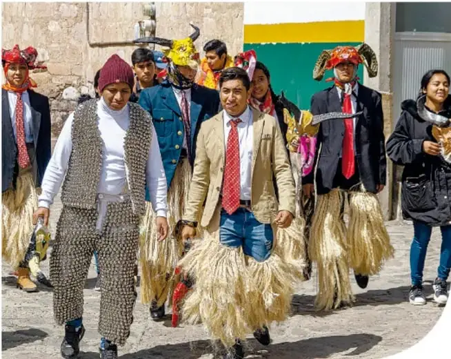 Fotografía de un danzante con máscara amarilla de cuernos negros, barba, saco azul, camisa blanca y corbata roja, sosteniendo un látigo. Al fondo se ven papeles de colores colgando.