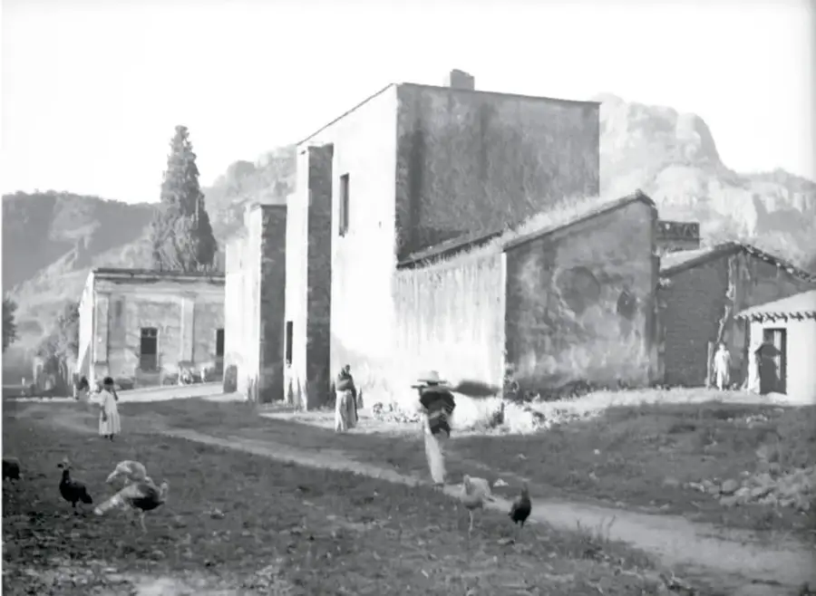 Fotografía antigua en blanco y negro de un caserío con muros altos y techos de terrado, montañas al fondo y personas con animales domésticos en el camino.