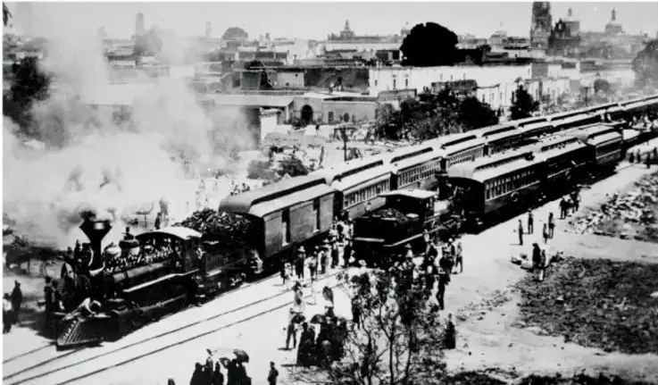 Fotografía en blanco y negro de una locomotora de vapor con varios vagones de pasajeros detenida en una estación; numerosas personas caminan alrededor y al fondo se observan edificios de una ciudad.