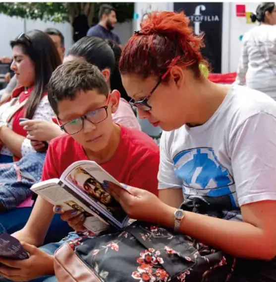Fotografía de una madre y un niño leyendo juntos un libro en la calle, entre otras personas.