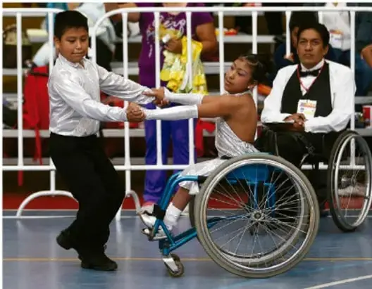 Fotografía de un evento: un niño de pie ayudando a una niña en silla de ruedas, y un hombre en silla de ruedas con chaleco y pase de acceso, en un espacio con rejas y público de fondo.