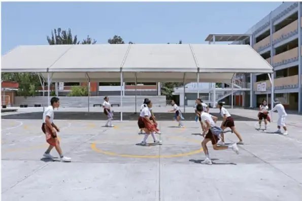 Fotografía de estudiantes con uniforme blanco y pantalón oscuro jugando basketball en una cancha al aire libre techada, con edificios escolares al fondo.