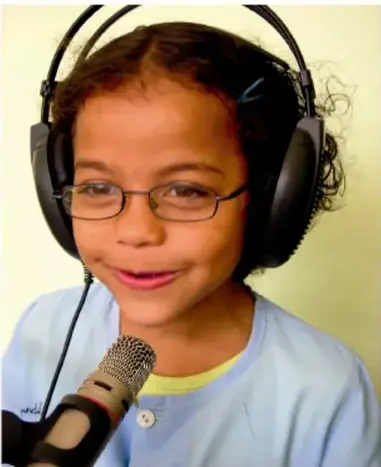 Niña con audífonos grandes y micrófono sonríe mientras graba, con fondo amarillo.