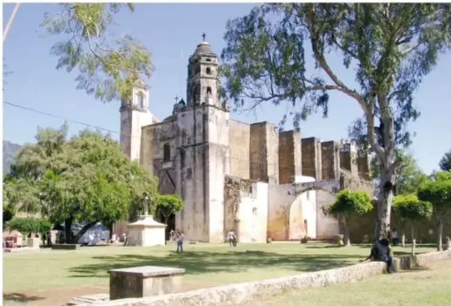 Fotografía del templo y exconvento de la Natividad de Tepoztlán, con su fachada de cantera, campanario y un parque con árboles al frente.