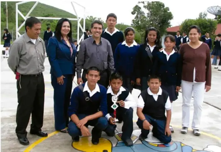 Fotografía de un grupo de estudiantes de telesecundaria con sus maestros en la cancha de la escuela, durante el evento de clausura; algunos estudiantes sostienen un trofeo.