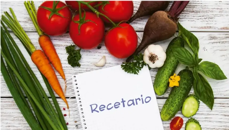 Fotografía de verduras frescas (zanahorias, jitomates, cebollín, betabel, pepinos, ajo, hierbas) sobre una mesa de madera, junto a un cuaderno con la palabra 'Recetario'.