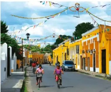Calle de un pueblo con personas caminando y andando en bicicleta, decorada con banderines, mostrando vida comunitaria.