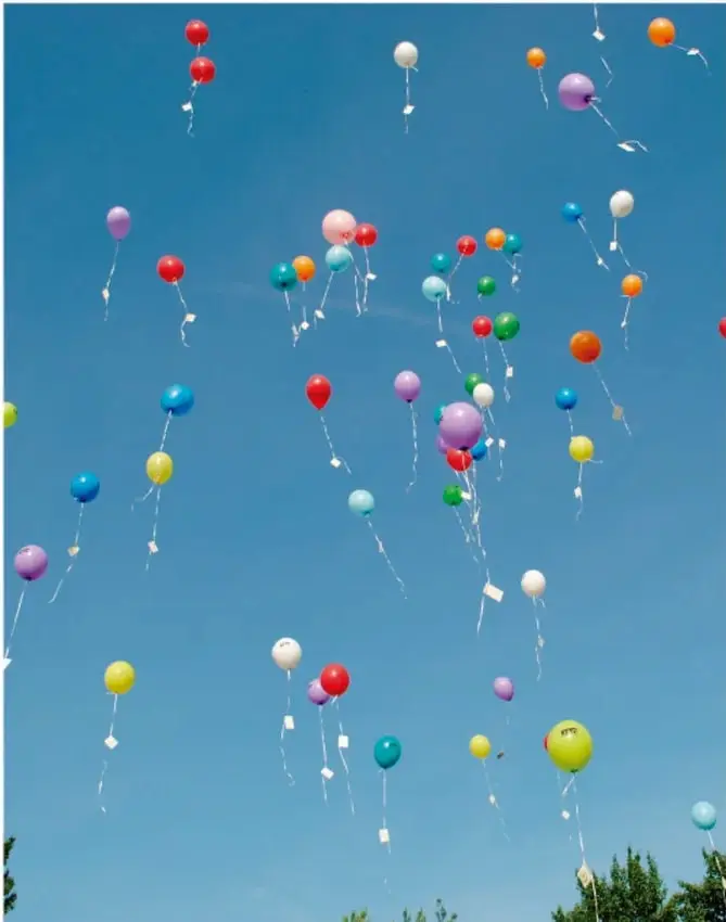 Fotografía de numerosos globos de colores (rojo, azul, amarillo, verde, morado, blanco) elevándose en un cielo azul despejado, con pequeñas etiquetas blancas colgando de sus hilos.