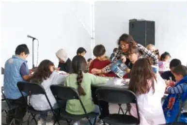 Fotografía de un grupo de estudiantes y una maestra organizando materiales en una mesa dentro del aula.