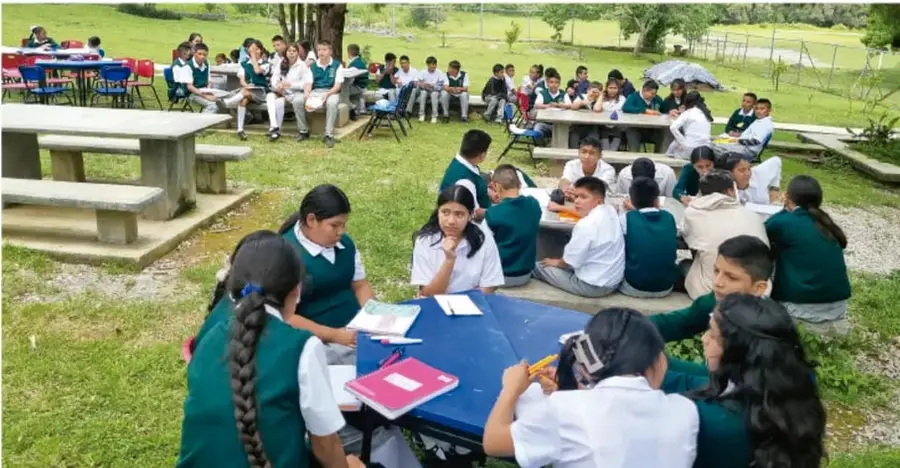Fotografía de estudiantes de Telesecundaria trabajando en equipos al aire libre, sentados en mesas y círculos sobre el pasto, con cuadernos, carpas y materiales escolares.
