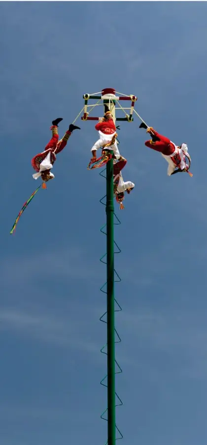 Fotografía de los voladores de Papantla: cuatro danzantes colgados de cuerdas en la parte alta de un mástil verde, girando boca abajo sobre un cielo azul, con traje rojo y blanco tradicional.