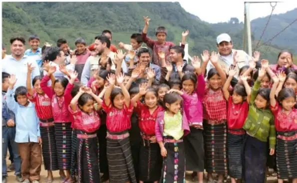 Fotografía de un grupo numeroso de niñas, niños y adultos de una comunidad indígena de montaña que saludan con las manos alzadas, vestidos con ropa tradicional colorida.