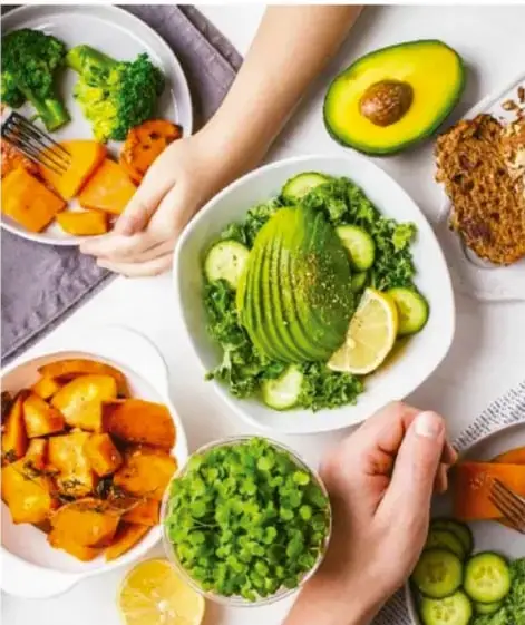Fotografía de varias manos sirviendo alimentos saludables (brócoli, aguacate, garbanzos, camote, ensaladas) en platos sobre una mesa.