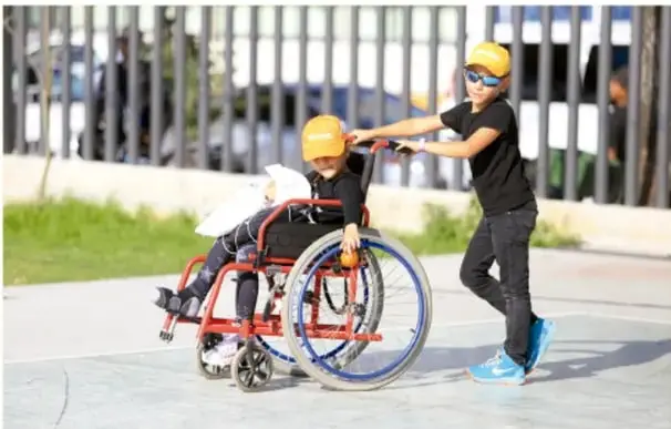 Fotografía de dos niños con casco amarillo jugando al airear: uno empuja a otro que va en una silla de ruedas sobre una cancha, ilustrando la inclusión y el compañerismo.