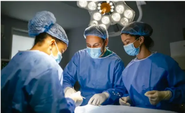 Fotografía de tres profesionales de la salud (quirófanos) con ropa azul de cirugía, mascarillas y gorros, trabajando juntos en un procedimiento; ilustra el trabajo colaborativo y las metas compartidas.