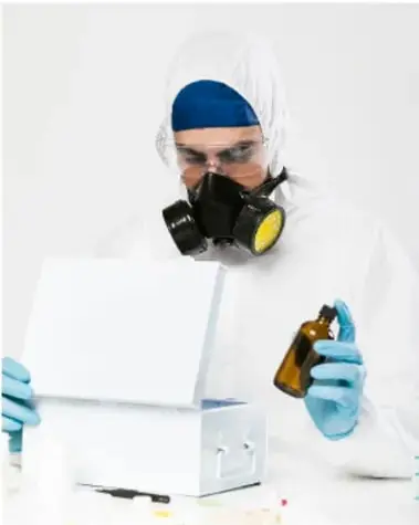 Persona con traje blanco de protección completa, cubrebocas respirador con filtros amarillos, gafas de seguridad y guantes azules, sosteniendo un frasco ámbar sobre una caja blanca.