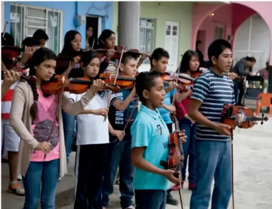 Fotografía de niñas, niños y jóvenes tocando violines y otros instrumentos de cuerda en una banda escolar al aire libre, frente a un edificio de colores.