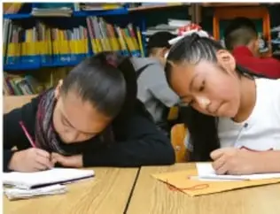 Dos niñas escriben en sus cuadernos en un salón con estantes de libros.