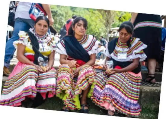 Tres mujeres con vestimenta tradicional de colores brillantes y plisados, sentadas en un espacio abierto.