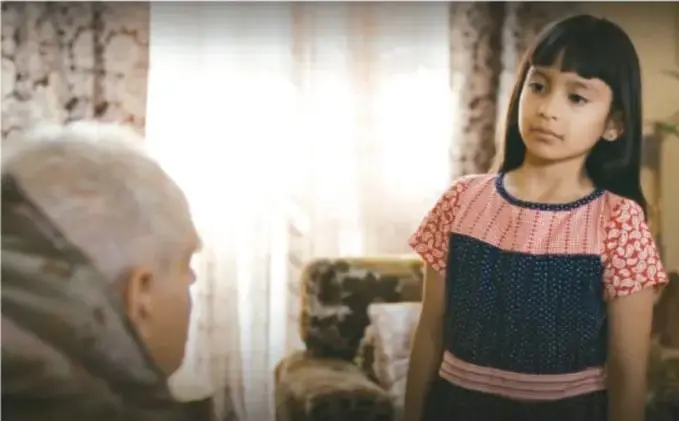 Fotograma del cortometraje: una niña de cabello largo con blusa de mangas cortas observa a una persona mayor sentada, en un interior con cortinas y sillón.