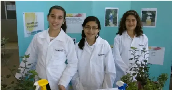 Fotografía de tres adolescentes con bata de laboratorio en una feria de ciencias, junto a un proyecto con plantas y carteles.