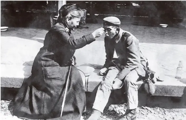 Fotografía en blanco y negro de una mujer uniformada que ayuda a un soldado herido o cansado, sentado en un muro; escena de la Segunda Guerra Mundial o del periodo de entreguerras.
