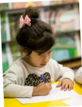 Fotografía de una niña escribiendo en una hoja sobre un escritorio, con una biblioteca de fondo.
