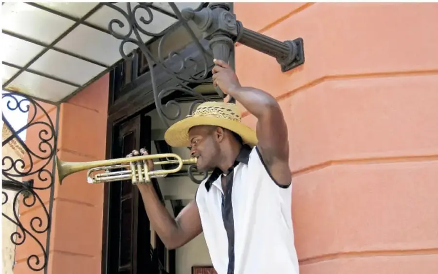 Fotografía de un músico afrodescendiente tocando una trompeta bajo un balcón con herrería colonial, con camisa blanca y sombrero de paja; representa la presencia viva de la música afroamericana en la actualidad.