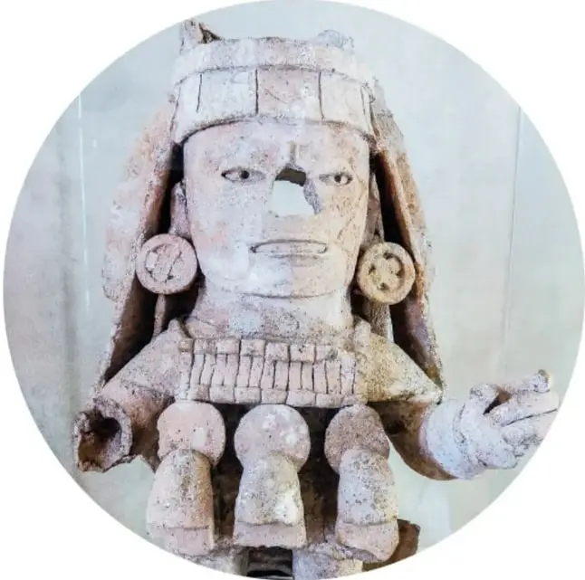 Escultura maya de piedra de un personaje con tocador o penacho, aretes circulares y collar; una de sus manos está extendida (piezas del Museo de Arqueología Maya).