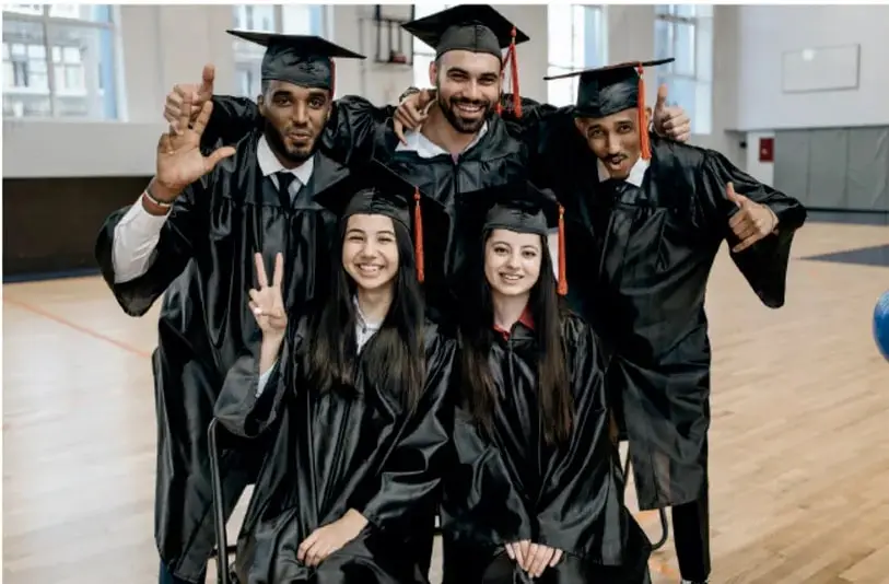 Fotografía de cinco jóvenes graduados con toga y birrete, posando sonrientes y haciendo gestos de celebración (pulgar arriba, señal de paz) en un gimnasio.