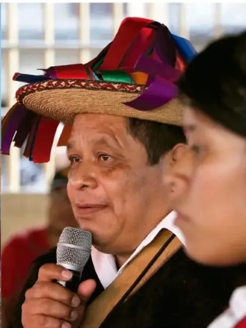 Fotografía de un hombre indígena con sombrero de paja adornado con cintas de colores, sosteniendo un micrófono, y una mujer de perfil junto a él; parece una asamblea o evento comunitario.