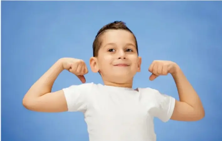 Niño con playera blanca flexionando los brazos y apretando los puños, mostrando tensión en bíceps y antebrazos.