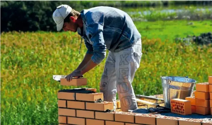 Fotografía de un albañil con gorra y overol construyendo un muro de ladrillo rojo junto a una cubeta y más ladrillos, en un campo verde.