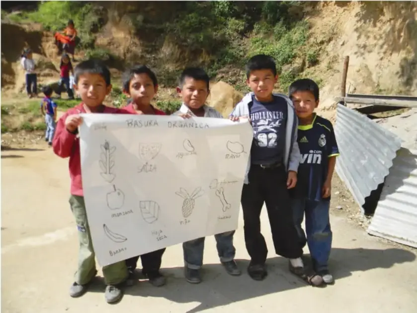 Fotografía de cinco niños de primaria sosteniendo un cartel titulado 'BASURA ORGÁNICA' con dibujos de residuos como manzana, plátano, cáscara de huevo, hueso, piña y hoja seca. Están al aire libre, en un camino de tierra con una ladera y una lámina de metal al fondo.