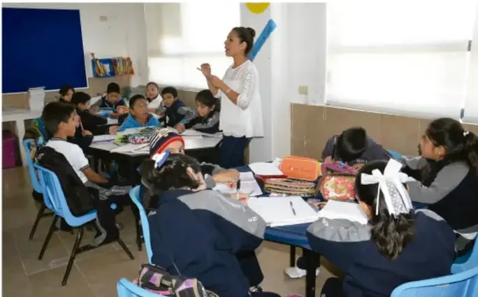Fotografía de un salón de clases: una maestra de pie dirige una actividad mientras los estudiantes, sentados en equipos, trabajan con cuadernos y materiales sobre las mesas.