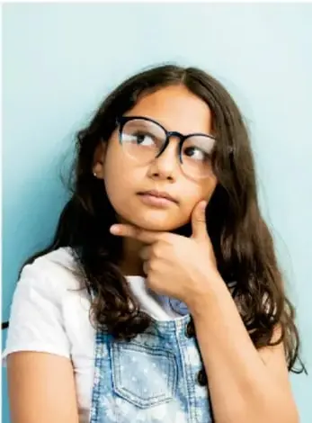 Niña pensativa con lentes y overol de mezclilla, apoyada en la mano.