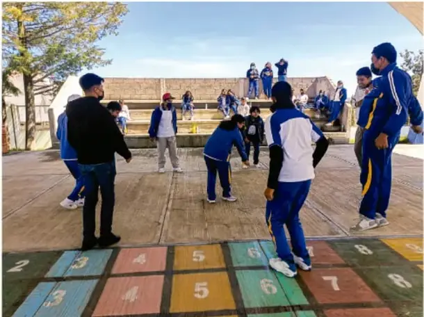 Fotografía de estudiantes con uniforme azul jugando en el patio escolar sobre un petate con números pintados; otros esperan su turno en gradas. Crédito: Luis Eduardo García Nietel, INAI/TAILA.