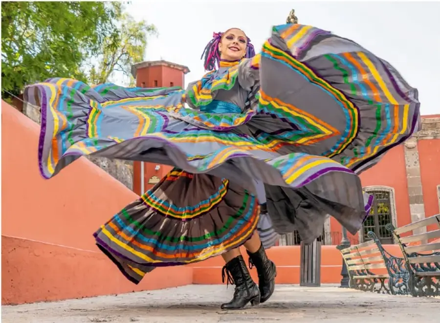 Fotografía de una bailarina con vestido folclórico de colores (gris con listones morados, verdes y amarillos) girando en una plaza con edificios coloniales y bancas de hierro al fondo.
