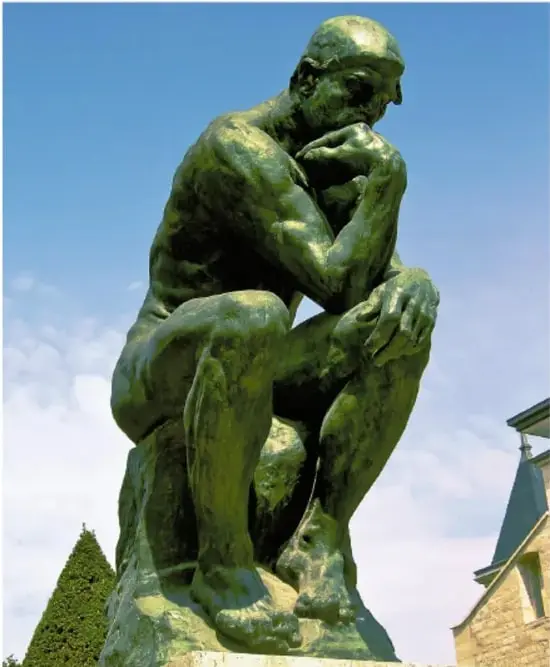 Escultura de bronce de El Pensador de Auguste Rodin: un hombre sentado, encorvado, con el mentón apoyado en la mano en actitud de profunda reflexión, sobre un peñasco, con cielo azul y vegetación al fondo.