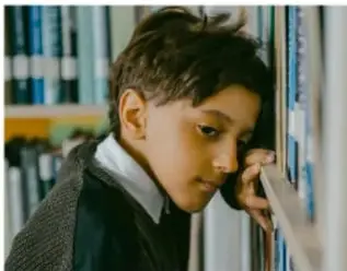 Niño con expresión de tristeza o preocupación, apoyado en una pared, con libros al fondo.