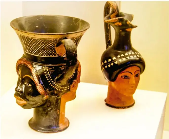 Fotografía de dos vasijas cerámicas antiguas de figuras humanas en vitrina de museo: una copa cuyo pie tiene forma de cabeza de persona negra y una botella con rostro femenino, ejemplo de representaciones de la antigüedad ligadas a la discriminación.